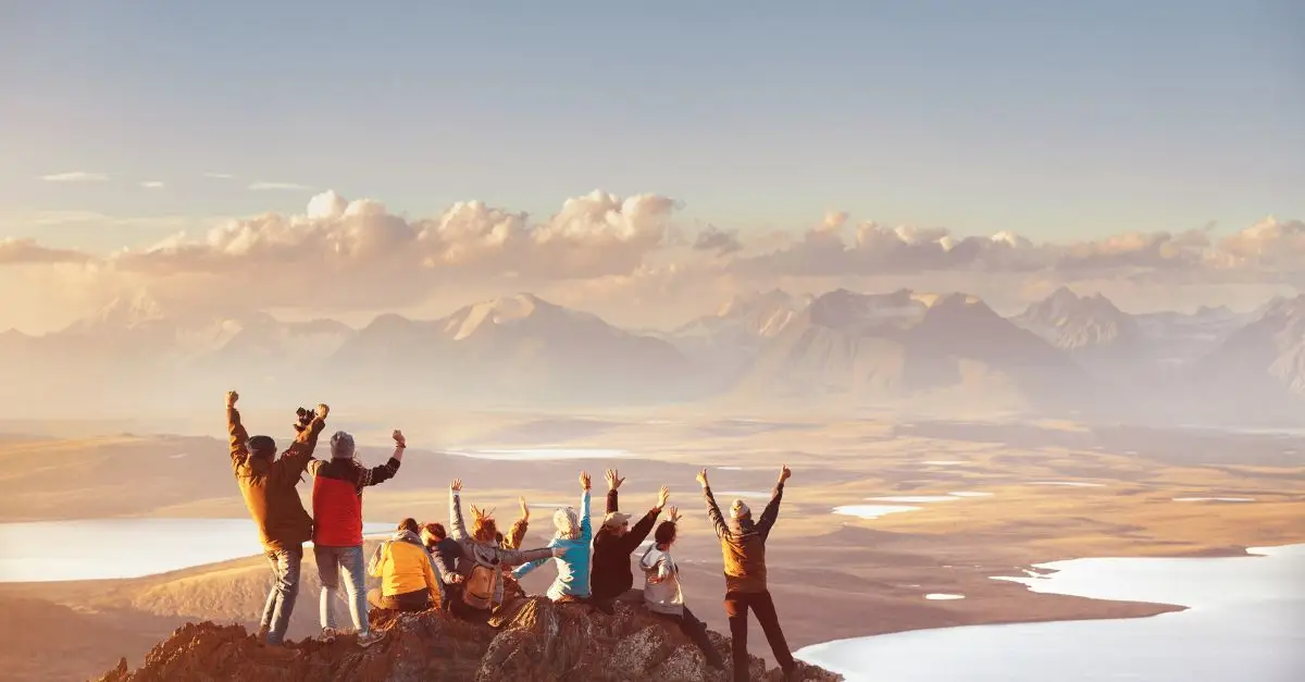 A group of people standing on a mountain summit overlooking a vast landscape, celebrating together and representing the power of diverse strengths working toward a shared goal.