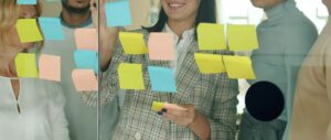 A diverse team gathered around a glass wall covered with sticky notes, working through ideas and different perspectives to solve a shared challenge.