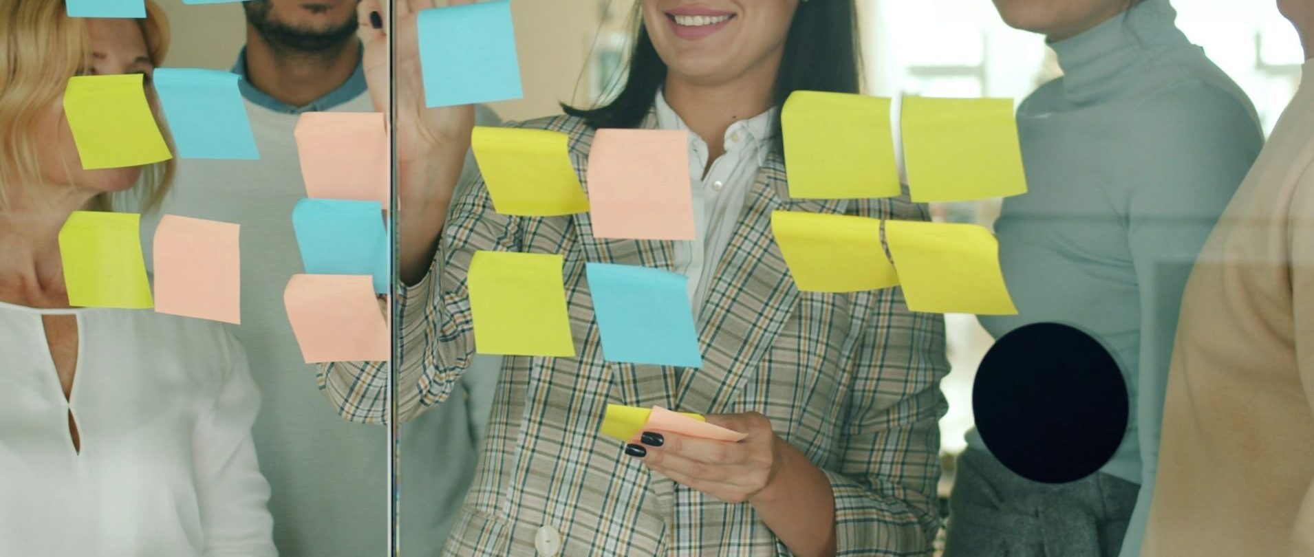A diverse team gathered around a glass wall covered with sticky notes, working through ideas and different perspectives to solve a shared challenge.